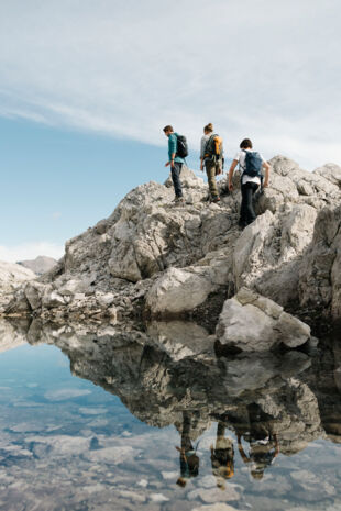 Drei Wanderer gehen entlang eines kleinen Gebirgssees durch karges Gebirge