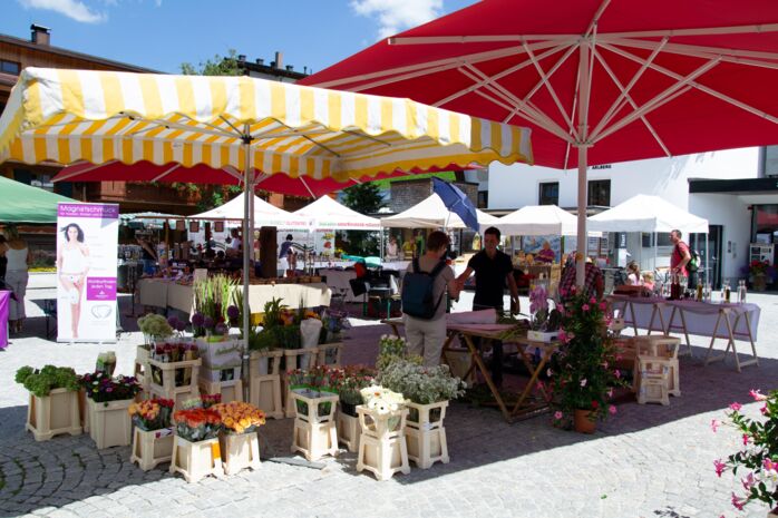 Tannbergermarkt im Freien mit bunten Blumen, Verkaufsständen und großen Sonnenschirmen an einem sonnigen Sommertag.