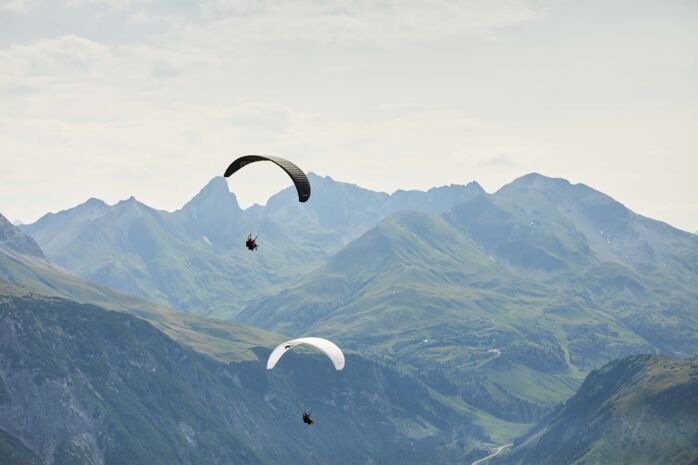 Zwei Paragleiter von hinten zu sehen und im Hintergrund eine große weite zu sehen mit Berggipfeln