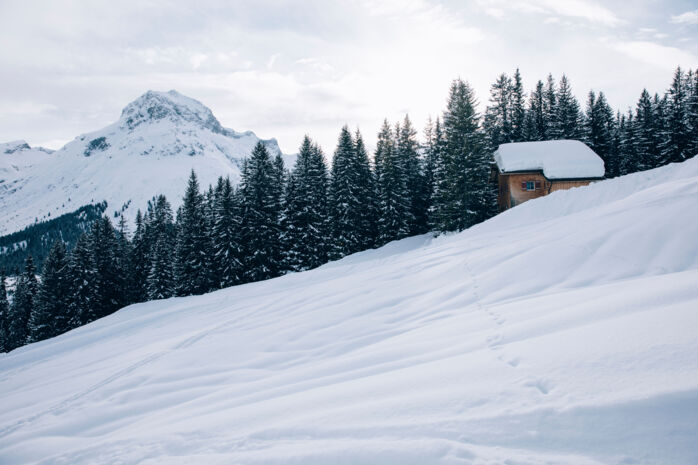Blick auf eine unbefahrene Tiefschneeabfahrt, im Hintergrund sieht man eine kleine Holzhütte vor dem traumhaften Bergpanorama