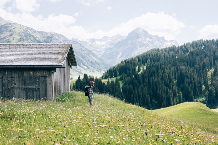 inmitten der blumigen Bergwiese steht ein Mann neben einer alten Holzhütte, im Hintergrund ist das Bergpanorama zu sehen