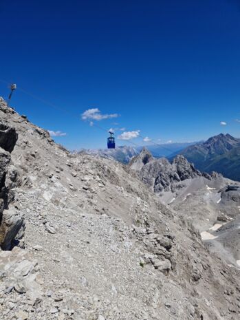 eine Gondel fährt über die felsige Berglandschaft, im Hintergrund sieht man das Bergpanorama