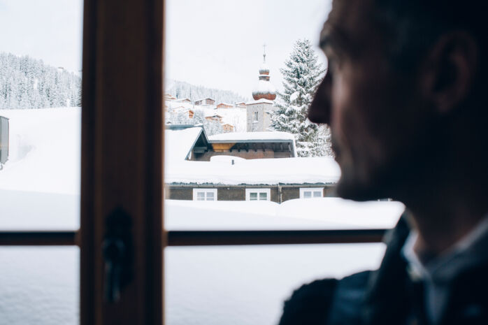 ein Mann blickt aus dem Fenster auf die schneebedeckten Dächer von Lech, im Hintergrund sieht man die winterlichen Wälder am Barg
