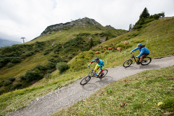 Zwei Radfahrer fahren einen Schotterweg im Gebirge hinab. Im Hintergrund erstrecken sich die weitläufigen Berge.