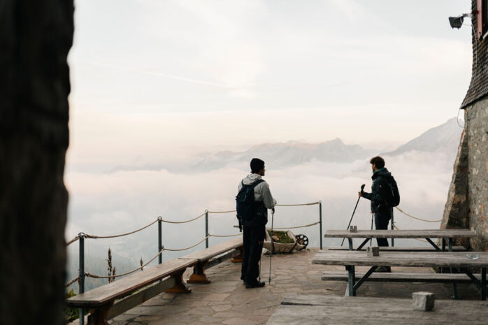 Zwei Wanderer stehen auf der Terrasse einer Hütte und blicken auf das weitläufige Bergpanorama und die tieferliegenden Wolken.