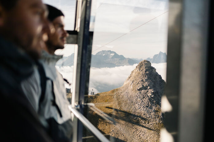 Zwei Wanderer blicken aus der Gondel hinaus auf die umliegenden Berge