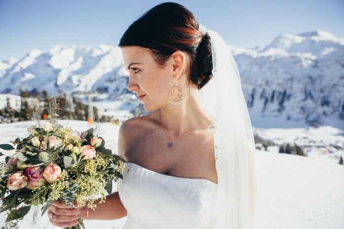 Die Braut hält ihren Hochzeitsstrauß und steht vor der winterlichen Bergkulisse bei ihrer Winterhochzeit am Arlberg