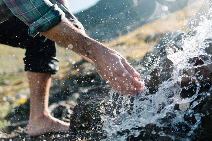 Ein Mann steht barfuß in einem Bach und berührt mit den Händen das klare Bergwasser