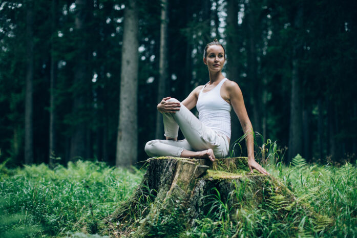 Frau sitzt im Wald auf einem Baumstumpf und macht Yoga Übungen