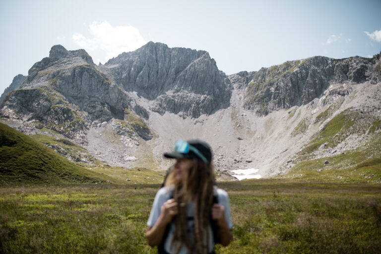 Wanderin mit Rucksack im Vordergrund unscharf, dahinter scharf im Fokus ein schroffes Bergmassiv mit Geröllhalde und Schneeresten.