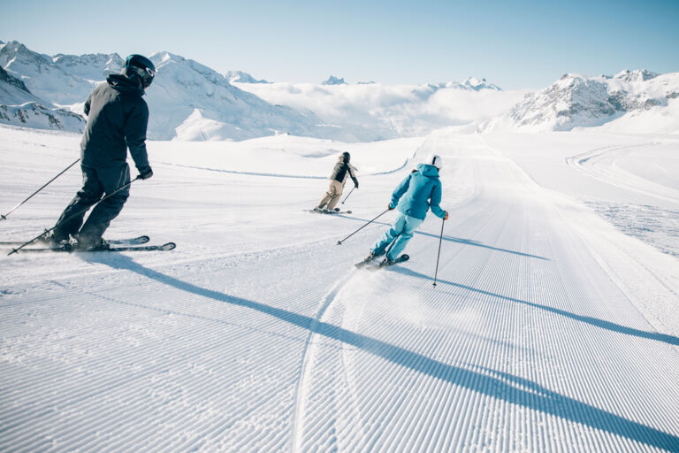 drei Skifahrer fahren bei klarem Sonneschein über die frisch präparierte Piste, mit traumhaften Bergkulissen im Hintergrund