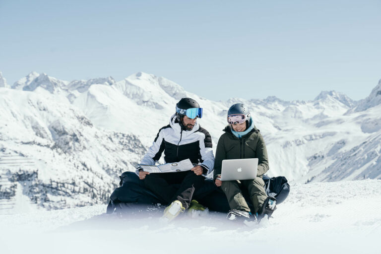 zwei Skifahrer sitzen am Berg mit ihrem Macbook während man im Hintergrund die schneebedeckten Bergspitzen sieht