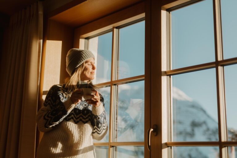 Frau sieht aus dem Fenster mit Teetasse in der Hand und lässt sich die Sonne auf das Gesicht scheinen