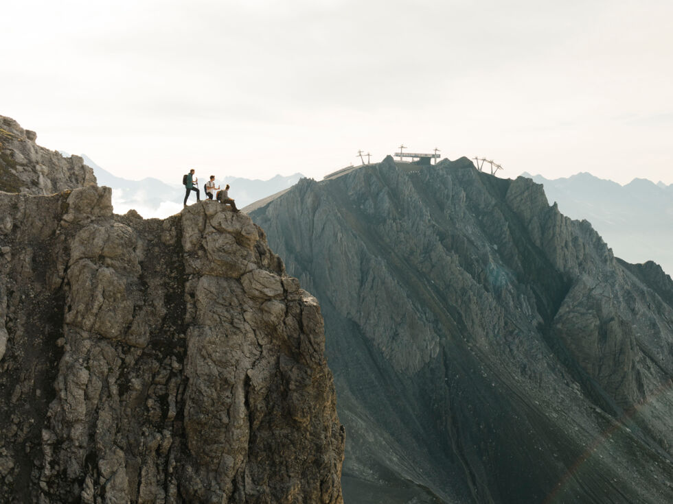 Wanderer sitzen und stehen an einem abschüssigen Felsvorsprung; im Hintergrund befindet sich ein Gebirgshang mit der Bergstation einer Seilbahn