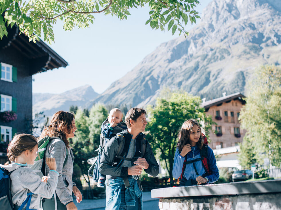 eine Familie steht vor dem Dorfbrunnen in Lech, im Hintergrund sieht man das Omeshorn