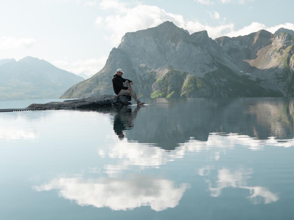 Mann sitzt auf einem Felsen an einem spiegelnden Bergsee vor Alpenkulisse.