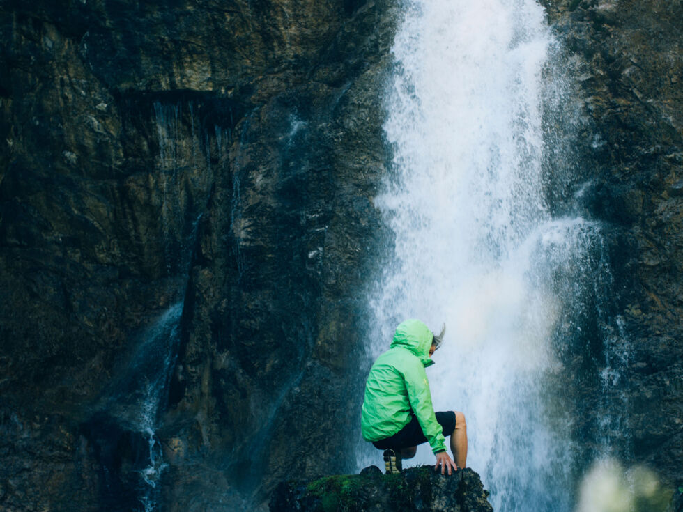 ein Kind sitzt auf einem Felsen und lauscht dem Plätschern des Wasserfalls, welcher  vor ihm zwischen den Felsen entspringt