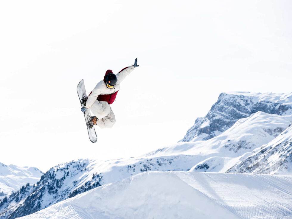 Freestyler springt über eine Schanze und im Hintergrund ist strahlend blauer Himmel und eine wunderschöne Bergkulisse zu sehen.