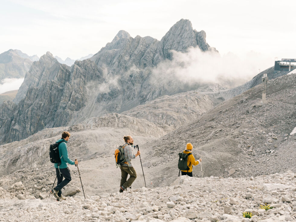 Drei Wanderer gehen auf einem felsigen Steig mit einem kargen Gebirgspanorama im Hintergrund.