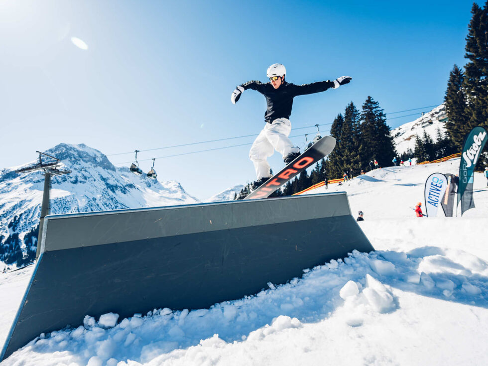 Freestyler fährt über eine Rail und im Hintergrund ist strahlend blauer Himmel und eine wunderschöne Bergkulisse zu sehen.