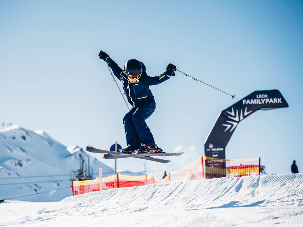 Junger Skifahrer springt mit gekreuzten Ski über einen Kicker im Lech Familypark, im Hintergrund Berge und blauer Himmel.