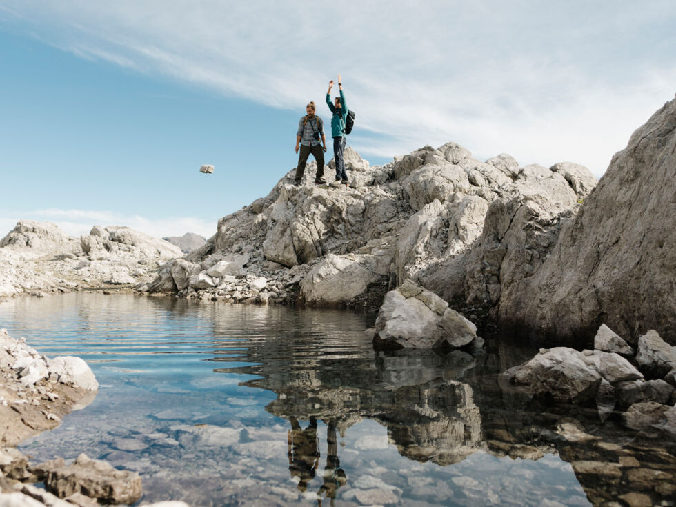 Zwei Wanderer stehen vor einen kleinem Gebirgssee und einer der beiden wirft einen Stein ins Wasser.